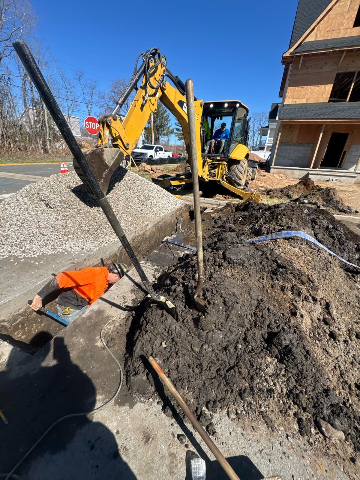 A backhoe digging along a curb at a new home site, crew member guiding the bucket.