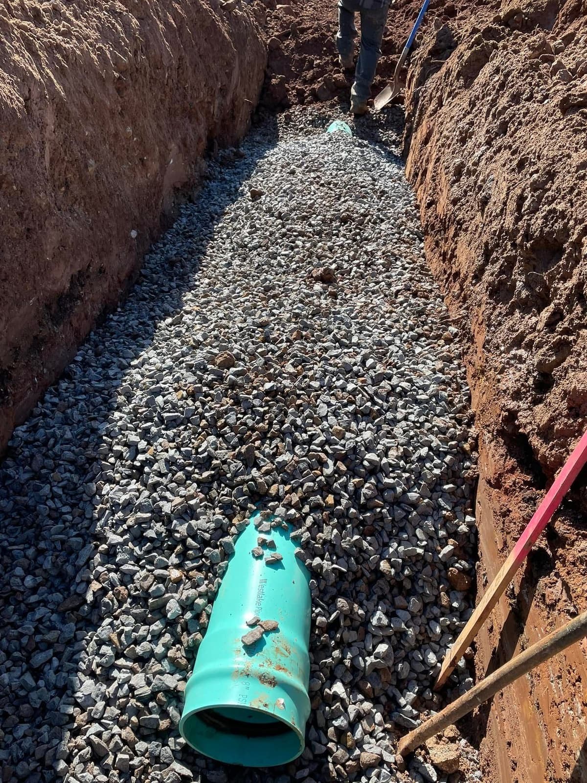 A deep drainage trench bedded with clean stone, with PVC pipe being laid in.