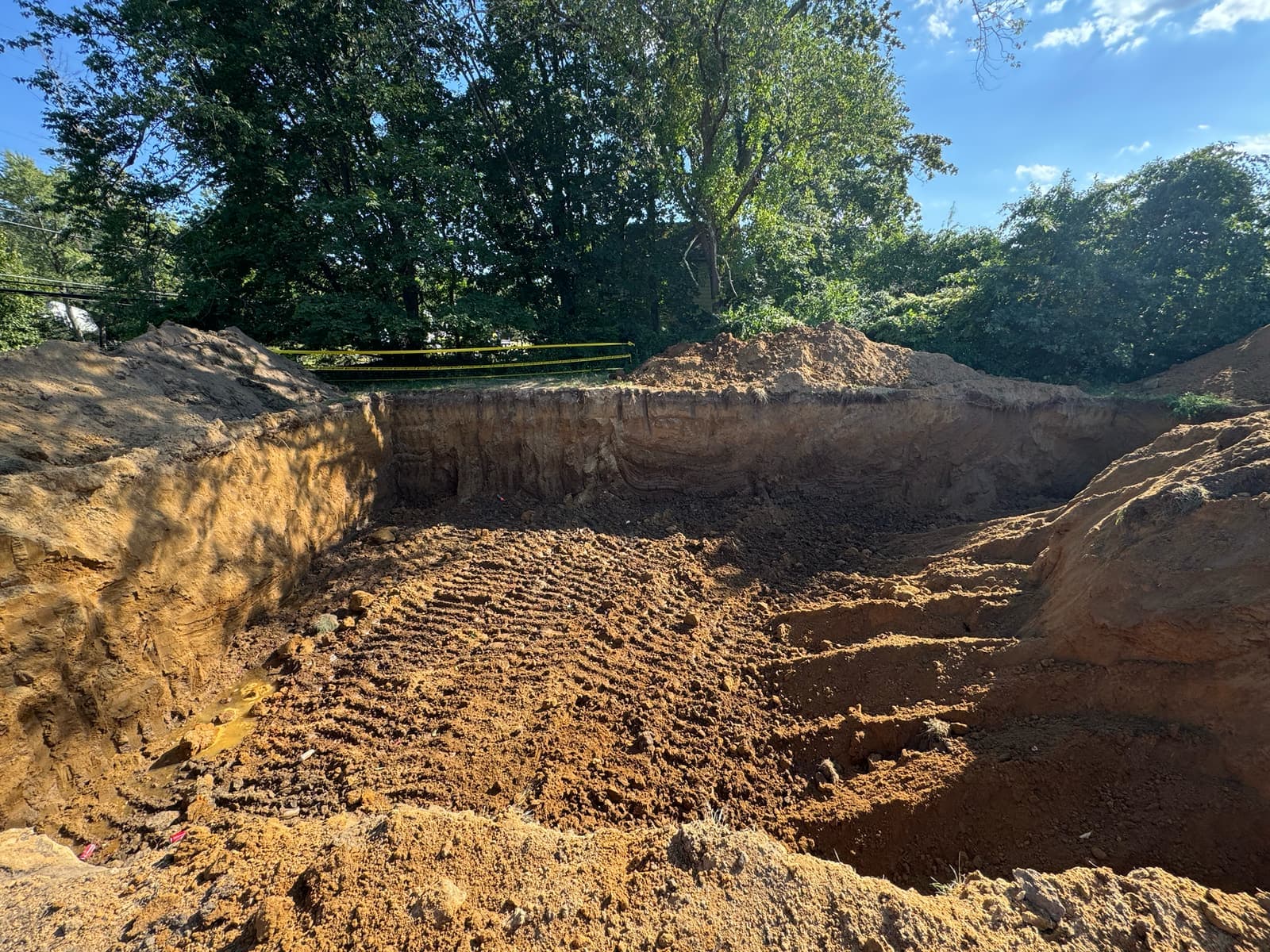A large, deep rectangular excavation in a wooded residential backyard.