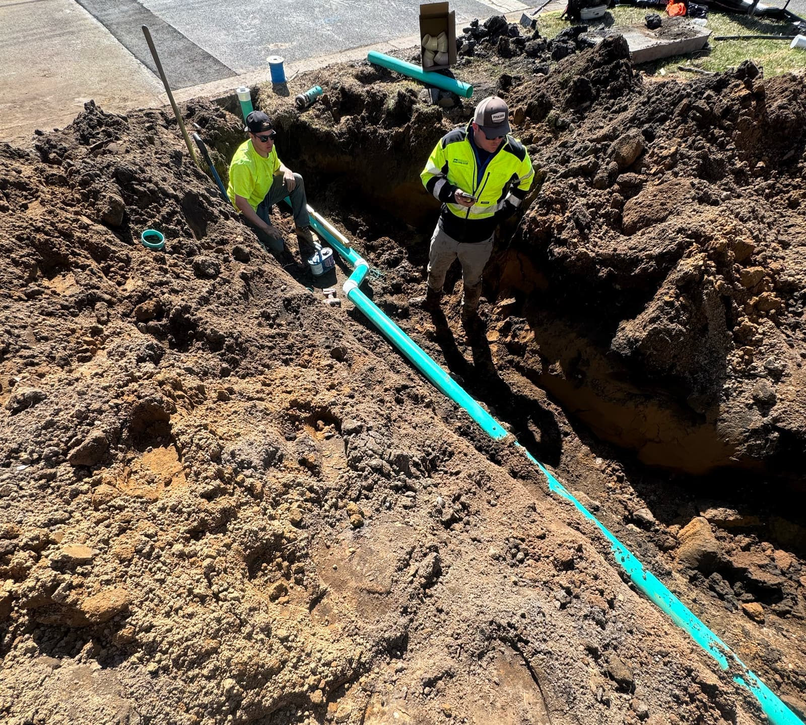 Two crew members laying green PVC pipe in an open trench next to a sidewalk.